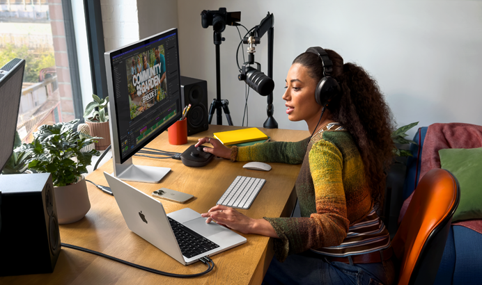 Sieviete savās mājās pie rakstāmgalda strādā ar MacBook Pro, kas savienots ar diviem ārējiem displejiem, – viņa ir uzlikusi austiņas un runā mikrofonā