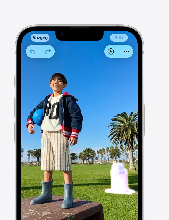 iPhone ekranında bir kişinin gülümseyerek sahilde çekildiği fotoğraf görünüyor, Düzeltme aracı ile arka plandaki bir koninin nasıl kaldırılacağı gösteriliyor