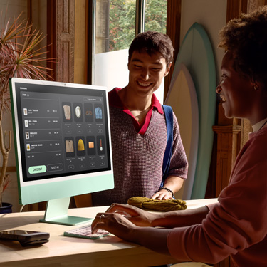 Eine Mitarbeiterin arbeitet hinter dem Tresen eines Ladens, der Surfbretter und andere Artikel verkauft. Sie schliesst mit einem iMac einen Einkauf für einen Kunden auf der anderen Seite des Tresens ab.