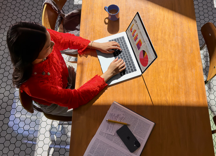 Eine Person, die an einem Tisch sitzt und ein MacBook benutzt, daneben eine Tasse, ein Smartphone und Papiere.