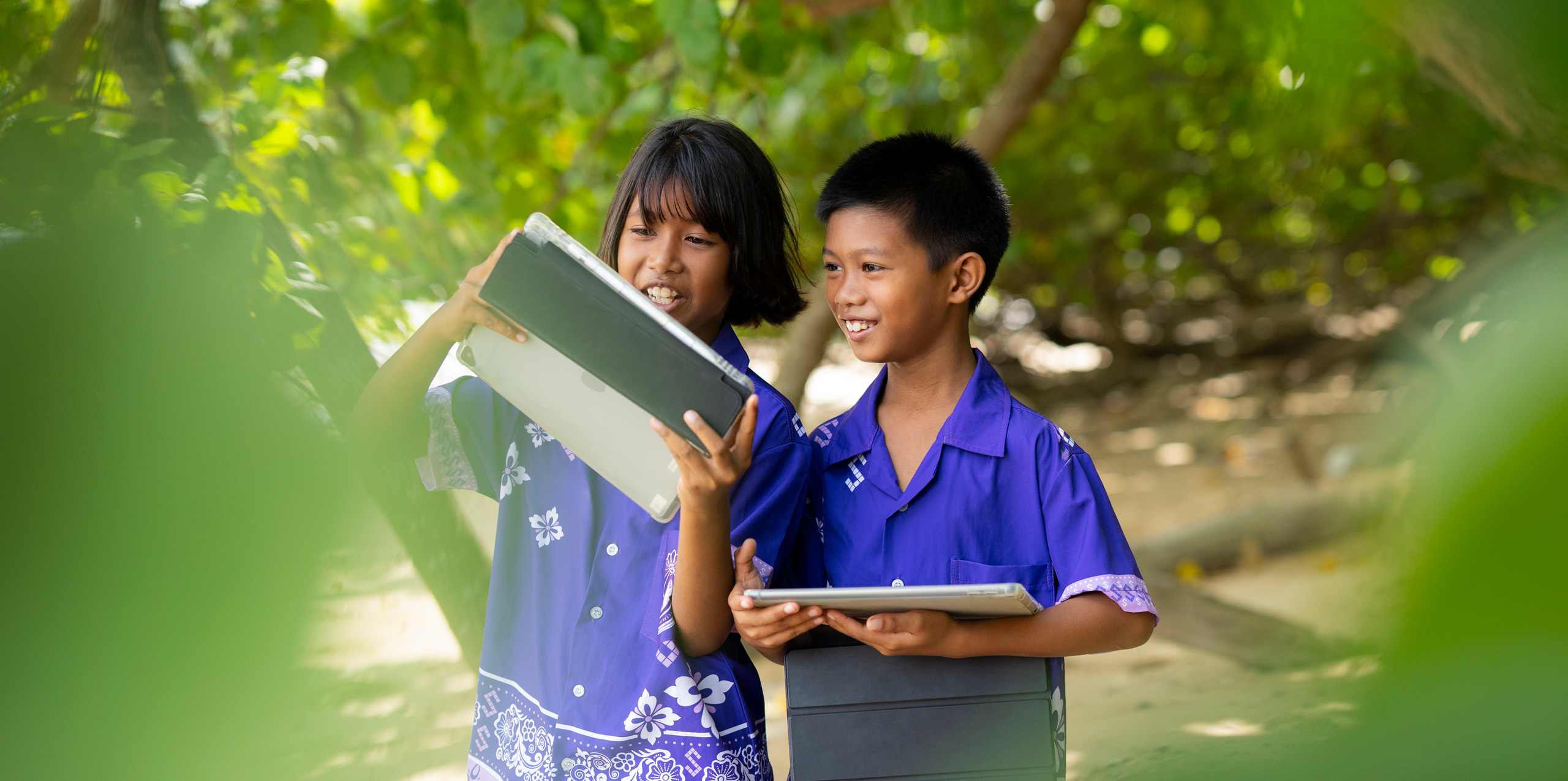 Two elementary students, one boy and one girl, standing outside amidst tropical trees, each holding an iPad. They are both looking at the girl’s iPad screen, smiling and engaged.