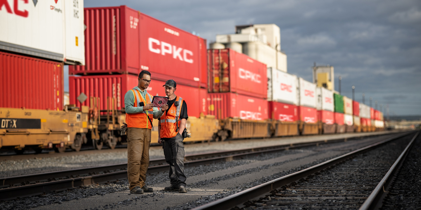 Two railroad workers reference an iPad while standing in a train yard beside a stationary train carrying shipping containers