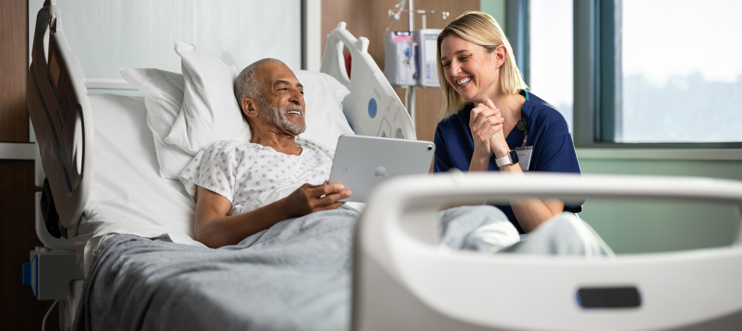 A male patient sitting in a hospital bed. He is smiling while looking at an iPad screen with a female nurse.